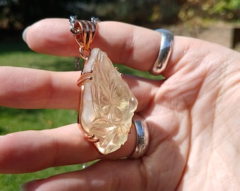 May include: A pendant necklace featuring a translucent, pale yellow carved gemstone with a floral pattern. The gemstone is secured in a rose gold-toned metal setting with prongs and hangs from a delicate silver-toned chain.