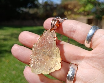 May include: A translucent, pale yellow carved gemstone pendant featuring a floral motif with detailed petals and leaves. The pendant is set in a rose gold-toned metal frame and hangs from a matching silver-toned metal chain necklace.