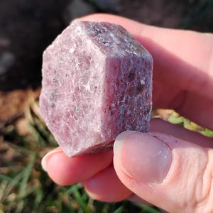 May include: A raw ruby gemstone with a pinkish-red colour and a faceted shape. The natural crystal has a rough, textured surface and is held in a hand. The image is taken in natural light, highlighting the stone's colour and form. The image text says "shot on moto g play - 2024 Valhalla Gems".