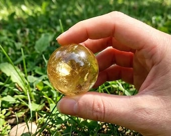 May include: A clear, golden-colored sphere held in a hand against a backdrop of green grass and foliage. The sphere appears to have internal golden inclusions, catching the sunlight. The scene is outdoors, with a natural, earthy aesthetic.