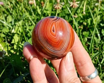 May include: A polished, spherical stone featuring concentric bands of deep red, burnt orange, and white. The smooth, rounded surface displays a natural layered pattern with a glossy finish, showcasing distinct circular rings throughout the orb.