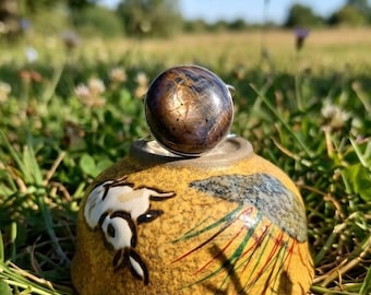 May include: A decorative bottle stopper with a round, dark-colored top and a yellow base. The base features a painted design of a rabbit and foliage. The stopper is set in a grassy field with a blurred background.
