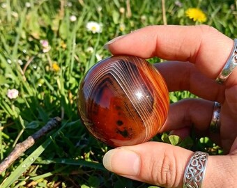 May include: A polished, spherical agate stone featuring deep red and orange hues with distinct, concentric white banding patterns. The surface is smooth and reflective, showcasing natural mineral layers that form a sharp, angular chevron design.
