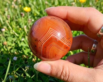 May include: A polished, spherical agate stone featuring vibrant orange and red concentric banding with subtle dark inclusions. The smooth, rounded surface displays a natural, intricate pattern of layered lines that form a distinct chevron design.