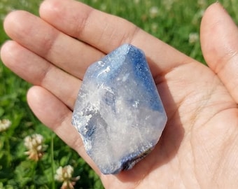 May include: A raw, irregularly shaped crystal specimen featuring a blend of translucent white and deep blue coloration. The stone has a faceted, crystalline texture with visible internal inclusions and a polished, smooth surface on its outer edges.