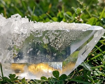 May include: A clear, transparent quartz crystal cluster featuring a jagged, crystalline top edge with a smooth, flat base. The mineral specimen displays natural internal fractures and facets that catch the light, creating a bright, reflective appearance.
