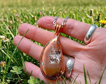 May include: A teardrop-shaped pendant featuring a polished brown and white stone set in a copper-toned metal frame, suspended from a silver-toned chain. The pendant is displayed alongside a plain silver-toned ring and a ring with an etched floral design.