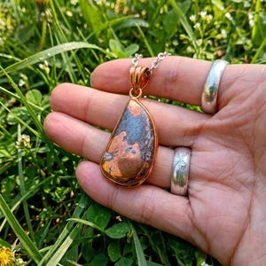 May include: A teardrop-shaped pendant featuring a polished copper and dark grey stone inlay, set in a copper frame and suspended from a silver-toned metal chain. The pendant displays a mottled texture with metallic copper and dark grey patches.