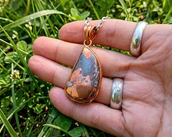 May include: A teardrop-shaped pendant featuring a polished copper and dark grey stone inlay, set in a copper frame and suspended from a silver-toned metal chain. The pendant displays a mottled texture with metallic copper and dark grey patches.