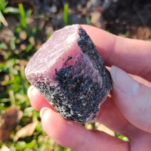 Puede incluir: Una gema rosa en bruto con matriz gris oscuro. La piedra se sostiene en una mano, con un fondo borroso de hierba verde y luz solar. El texto "shot on moto g play - 2024 Valhalla Gems" es visible en la parte inferior.