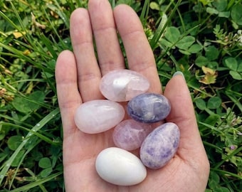 May include: A collection of six small, smooth, egg-shaped stones resting in an open palm. The stones feature a variety of colors and textures, including translucent pale pink, opaque white, and mottled shades of light purple and lavender.