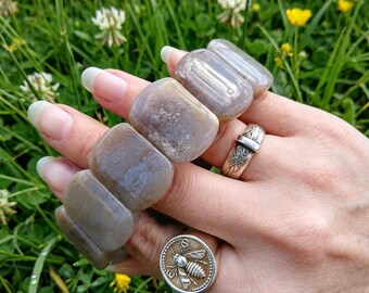 May include: A stretch bracelet composed of smooth, rectangular, translucent greyish-brown stone beads. The bracelet is worn alongside two silver rings: one featuring a detailed bee emblem on a circular face and another with an engraved band design.