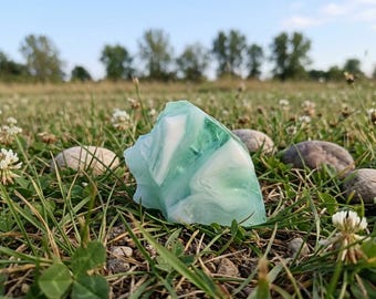May include: A translucent, light blue and white stone rests in a field of green grass, clover, and small rocks. The stone has a marbled appearance, with swirls of white and blue. The background features a blurred view of trees and a blue sky.