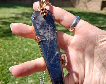 May include: A coffin-shaped pendant featuring a dark, polished stone set in a copper-toned metal frame. The design includes a small skull at the top and two skeletal hand accents gripping the sides of the stone. A thin metal chain is attached.