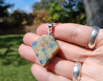 May include: A pendant featuring a rough, diamond-shaped stone with a tan, textured surface speckled with vibrant blue inclusions. The stone is secured by a silver-toned metal bail attached to a thin, matching silver-toned chain necklace.