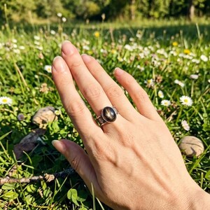 May include: A silver ring with an oval, black star sapphire gemstone. The ring is set in a detailed, ornate band. The hand is resting in a field of green grass and white flowers, with a blurred background of trees and sunlight.