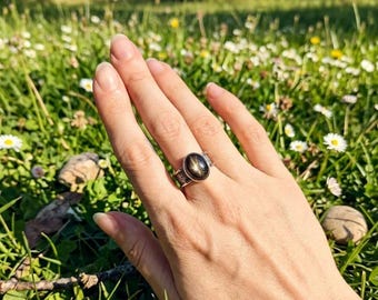 May include: A silver ring with an oval, black star sapphire gemstone. The ring is set in a detailed, ornate band. The hand is resting in a field of green grass and white flowers, with a blurred background of trees and sunlight.