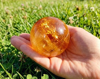 May include: A clear, golden-orange crystal ball held in an open hand, set against a backdrop of green grass and foliage. The sphere has internal golden inclusions, catching the sunlight. The scene is outdoors, with a blurred background of more greenery.