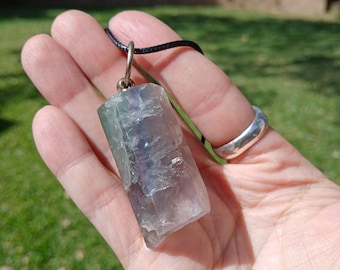May include: A raw, translucent fluorite crystal pendant featuring shades of purple, green, and clear mineral layers. The stone is secured by a silver-toned metal bail and hangs from a simple black cord necklace, showcasing natural geometric facets.