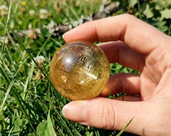 May include: A clear, golden-yellow crystal ball held in a hand against a backdrop of green grass and wildflowers. The sphere has internal inclusions and a polished surface, reflecting light. The image is taken outdoors in natural light.