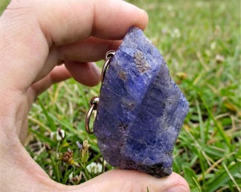 May include: A raw, deep purple-blue tanzanite crystal pendant featuring a rough, unpolished texture with natural facets and inclusions, securely attached to a simple, circular copper-toned metal jump ring for hanging or display purposes.