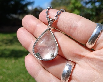 May include: A teardrop-shaped clear quartz pendant set in a silver-toned metal frame with a textured edge. The stone contains visible dark, star-shaped inclusions. The pendant hangs from a silver-toned metal chain link necklace.