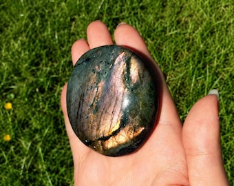 May include: A polished, circular labradorite stone featuring a dark green base with iridescent flashes of purple, gold, and blue light reflecting across its smooth, rounded surface, showcasing natural mineral inclusions and internal fractures.
