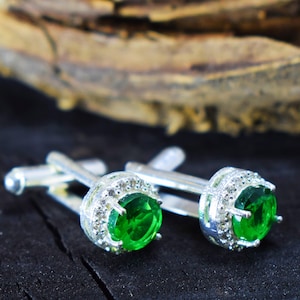 Puede incluir: Un par de gemelos plateados con una piedra central verde brillante de forma ovalada. Las piedras están rodeadas por un halo de piedras más pequeñas y transparentes. Se exhiben sobre una superficie oscura y texturizada.