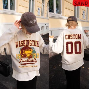 Puede incluir: Una sudadera de color arena con "WASHINGTON Football 1932" impreso en la parte delantera, con un gráfico de casco de fútbol. La parte trasera de la sudadera tiene impreso "CUSTOM 00". Se lleva una gorra marrón.