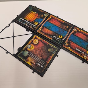 Op de afbeelding: Een bordspel met onderling verbonden tegels met kamerillustraties. De tegels bevatten "Ground Floor Staircase", "Hallway", "Entrance Hall" en "Armory". De spelstukken zijn zwart en de kamers zijn geïllustreerd met verschillende kleuren.