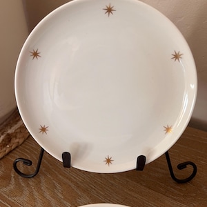 May include: White ceramic plate with gold starburst designs. The plate is displayed on a black metal stand. The plate has a simple, yet elegant design, perfect for serving food.