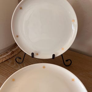 May include: Two white ceramic plates with gold starburst designs. The plates are displayed on a black metal stand, showcasing a minimalist aesthetic. The plates are on a wooden surface.