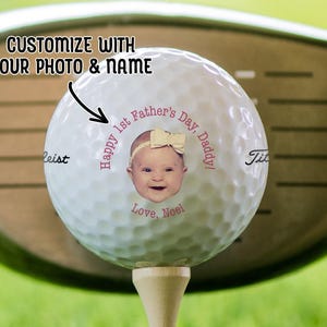 A golf ball on a tee with a golf club in the background. Golf ball has 1st Fathers Day photo design on it in the pink text color option.