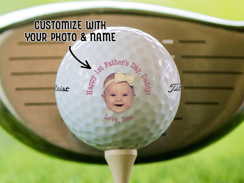 A golf ball on a tee with a golf club in the background. Golf ball has 1st Fathers Day photo design on it in the pink text color option.