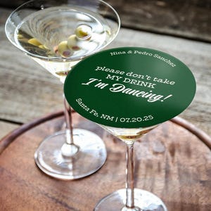 One coaster is being used as a drink cover on top of a martini glass on a wooden tray and table. Coaster says Please dont take my drink, Im dancing with wedding couples names, location, and wedding date.