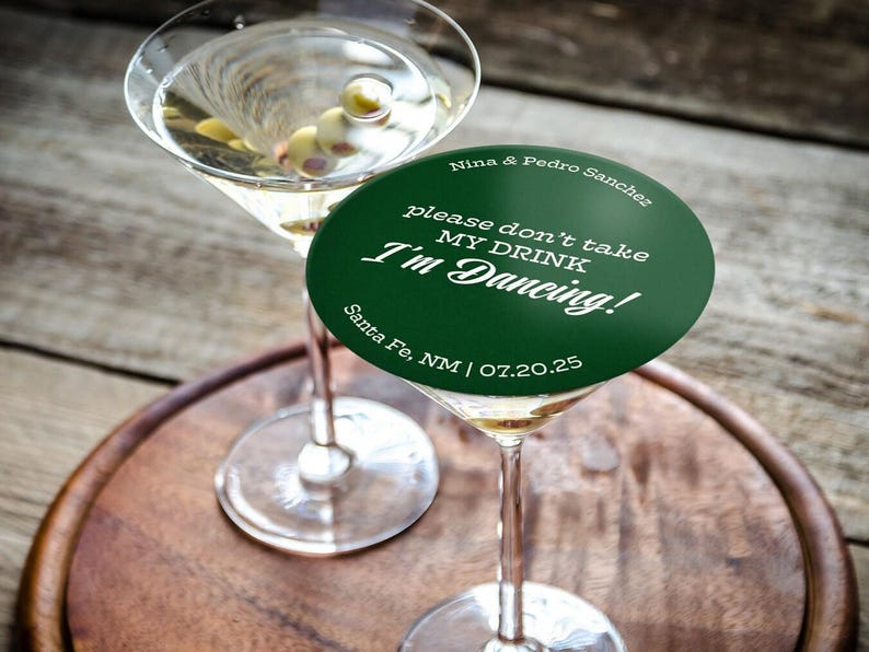 One coaster is being used as a drink cover on top of a martini glass on a wooden tray and table. Coaster says Please dont take my drink, Im dancing with wedding couples names, location, and wedding date.