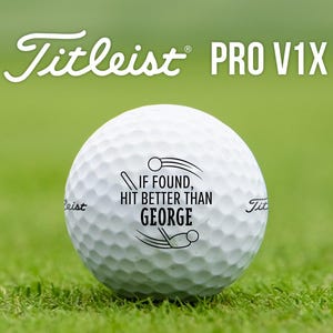 May include: A white golf ball with the text "IF FOUND, HIT BETTER THAN GEORGE" printed on it. The ball is sitting on green grass. The text "Titleist PRO V1X" is visible in the background.