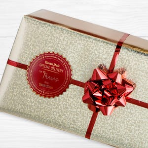 A gold foil cardstock Santa gift tag is shown on a Christmas present. The present is wrapped in gold paper and has a red bow and ribbon around it. The background of the picture is a white wood surface.