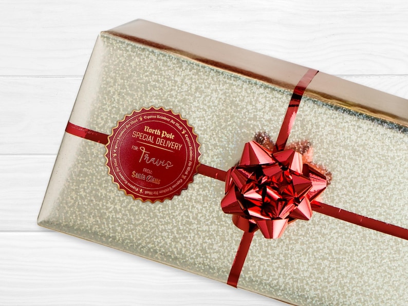 A gold foil cardstock Santa gift tag is shown on a Christmas present. The present is wrapped in gold paper and has a red bow and ribbon around it. The background of the picture is a white wood surface.