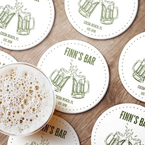 Multiple coasters sit on wooden surface with beer next to them.  Coasters feature bar design. This design uses green lettering on a white background, and a drawing of beer glasses. The name, established date, and location can be customized.