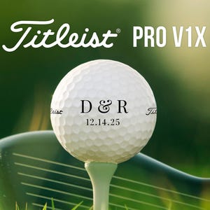 May include: A white Titleist Pro V1X golf ball with personalized text, including initials and a date, sits on a tee. A golf club is in the background. The image has a green, blurred background.