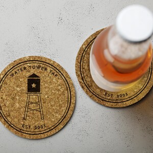 Two coasters sit on white surface with the words Water Tower Tap and an image of a water tower printed on it. One coaster has a drink on it.