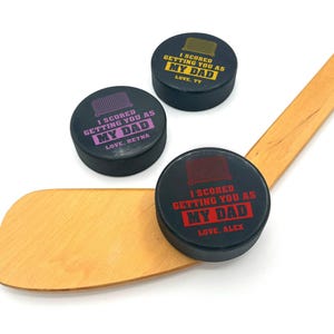 three hockey pucks with I scored you as my dad designs printed on them with the colors red, purple, & yellow with the names Alex, Reyna, and Ty