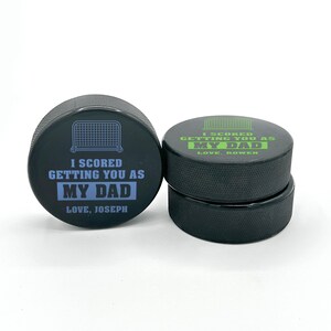 three stacked hockey pucks with printed  I scored you as my dad  designs in the color blue and green with the names Joseph and Rowen