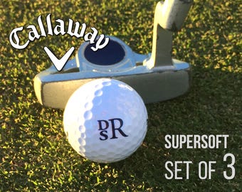 Può includere: Una pallina da golf bianca Callaway con le lettere DSR stampate in blu scuro, posizionata davanti a un putter in metallo argentato. Il testo SUPER SOFT SET OF 3 è visibile nell'angolo in basso a destra e il logo Callaway è in alto a sinistra.