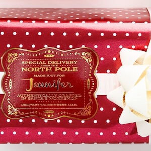 A gold foil cardstock Santa gift tag is shown on a Christmas present. The present paper is red with white polka dots. A white bow and ribbon are around the present. The box is seen on a white surface.