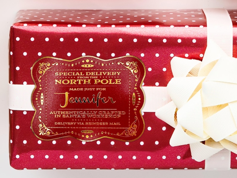 A gold foil cardstock Santa gift tag is shown on a Christmas present. The present paper is red with white polka dots. A white bow and ribbon are around the present. The box is seen on a white surface.
