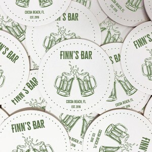 A stack of coasters are spread out across a surface. Coasters feature bar design. This design uses green lettering on a white background, and a drawing of beer glasses. The name, established date, and location can be customized.