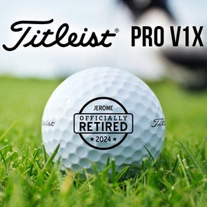 May include: A white golf ball with a black sticker that says "Jerome Officially Retired 2024" on it. The ball is sitting on green grass. The text "Titleist PRO V1X" is visible in the background.