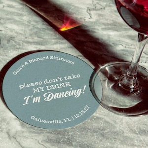 Coaster is shown with a wine glass on top of it and another off to the side. Coasters say Please dont take my drink, Im dancing with wedding couples names, location, and wedding date.